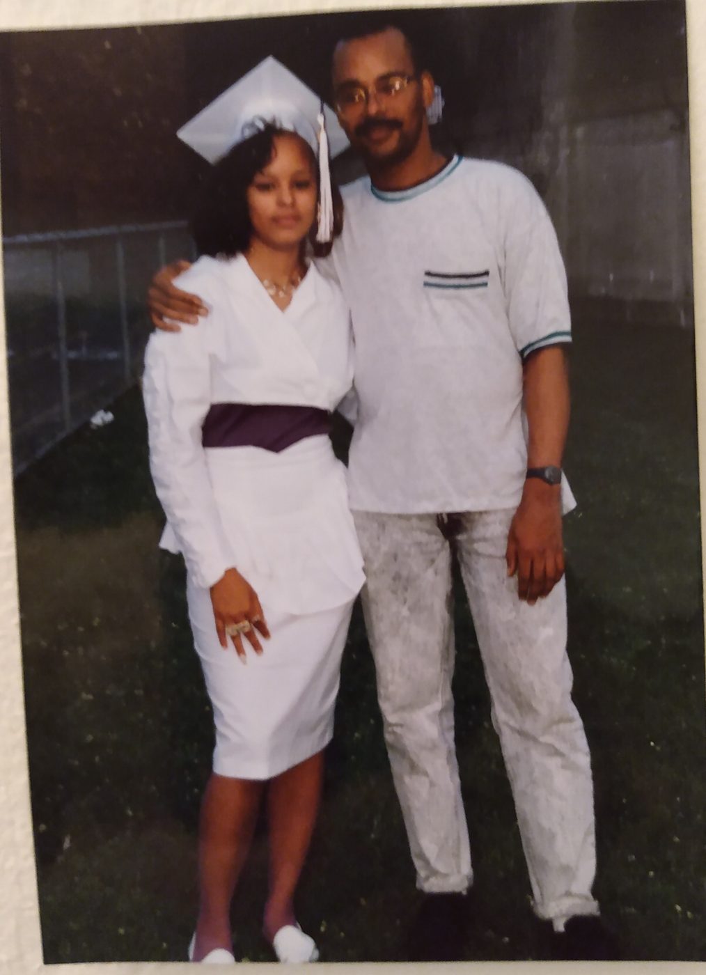 Sonya in a white cap and outfit on graduation day, with a family member